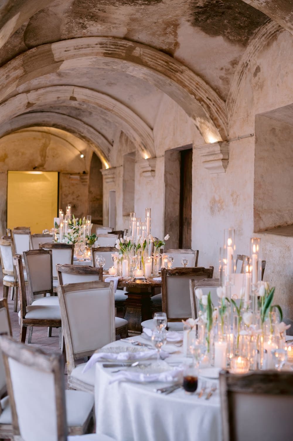 Vaulted cloister filled with candle-lit dining tables and white tulip arrangements — destination wedding dinner setting.