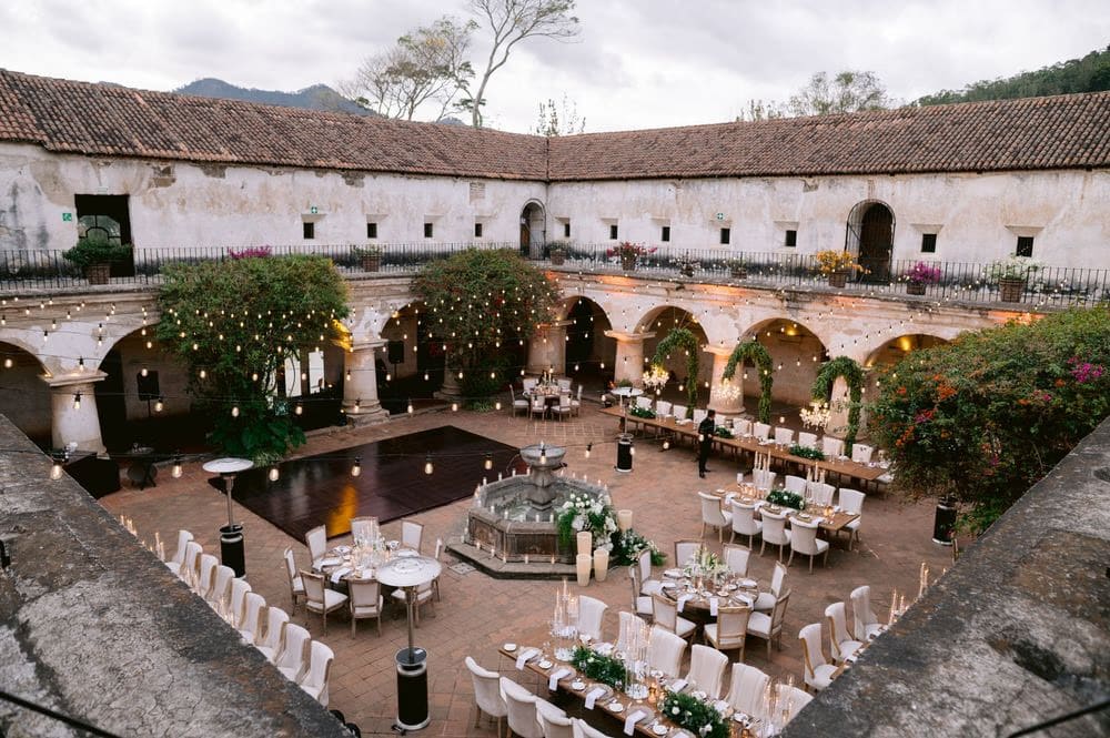 Wide overhead view of the candle-lit reception courtyard at Capuchinas Ruins, mountains rising behind the tile roofs.