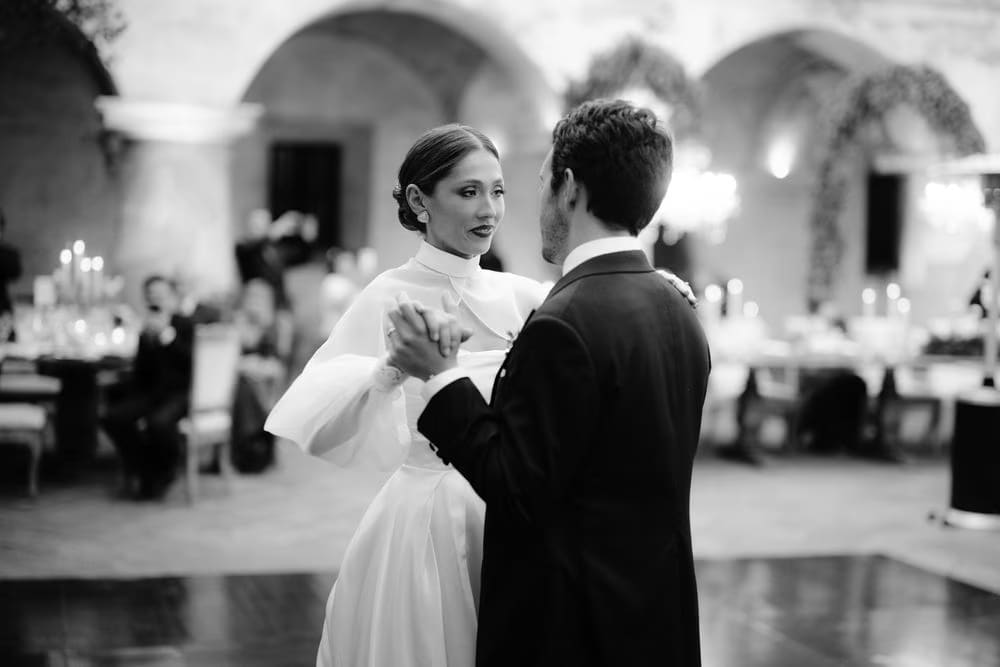 Black-and-white first dance — bride and groom hold each other on the dance floor as guests watch from candle-lit tables.