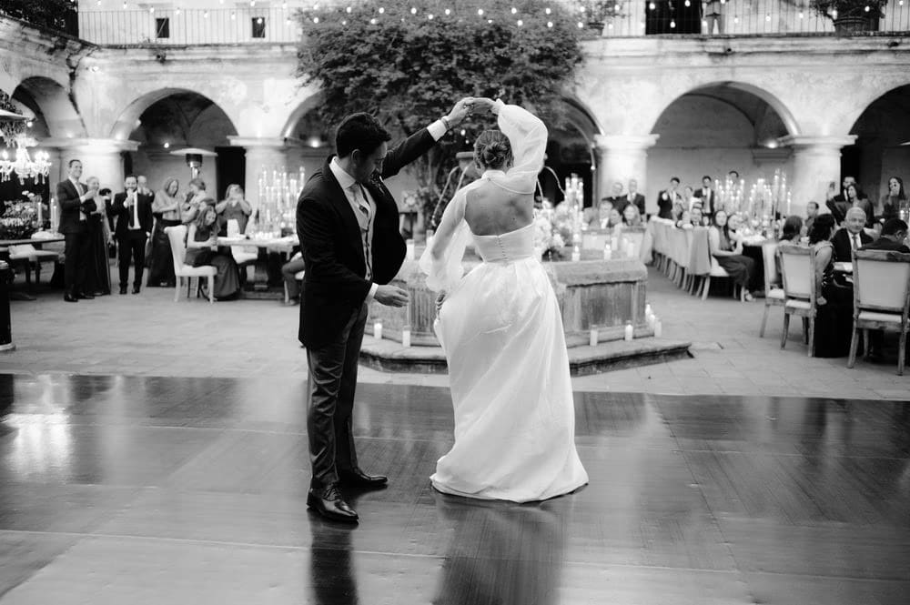 Groom twirls the bride on the wooden dance floor while guests applaud at the arcade-lined reception — black-and-white wedding dance.