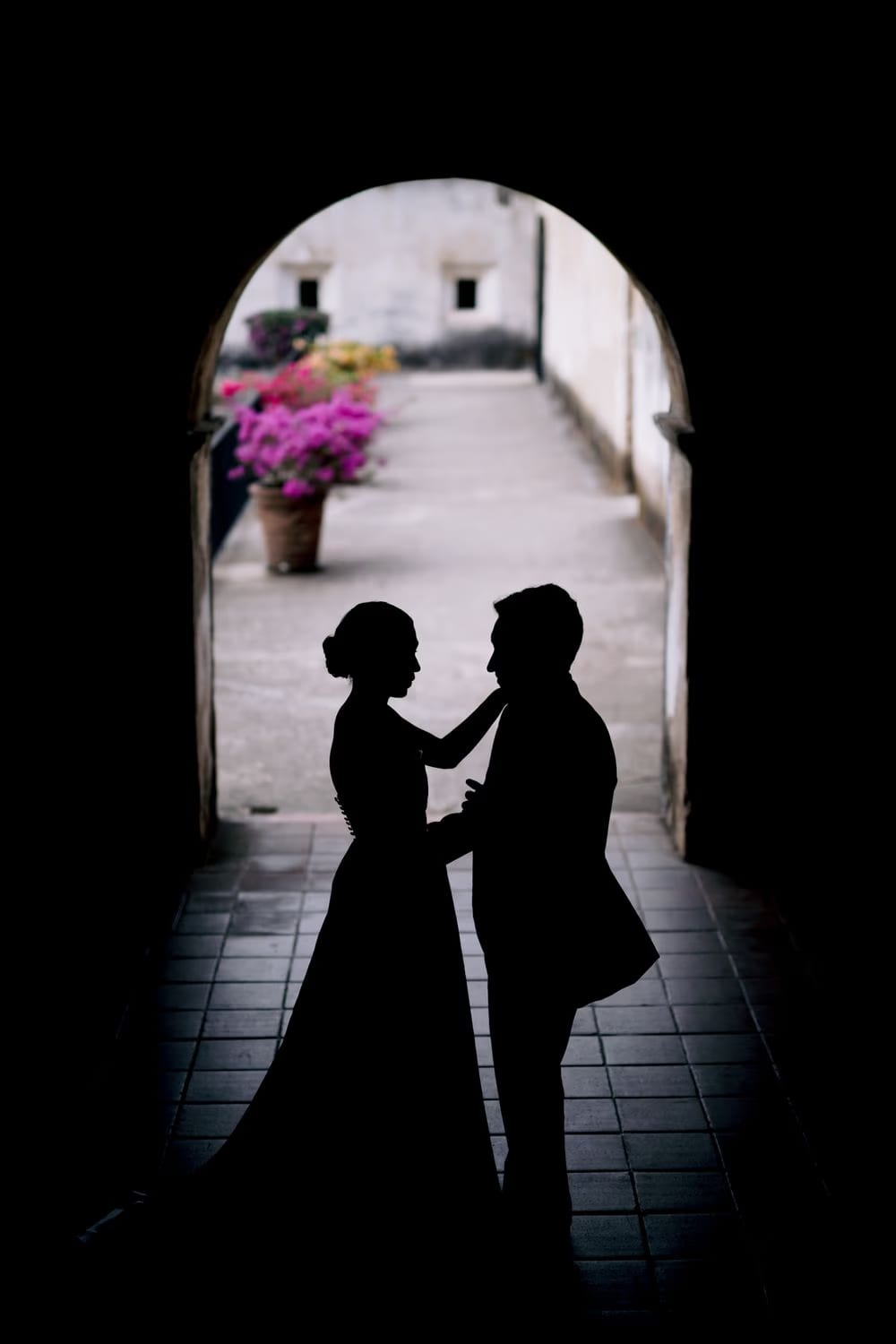 Silhouetted bride and groom face each other inside a stone archway at Capuchinas Ruins, bougainvillea blooming in the cloister beyond — destination wedding portrait in Antigua Guatemala.