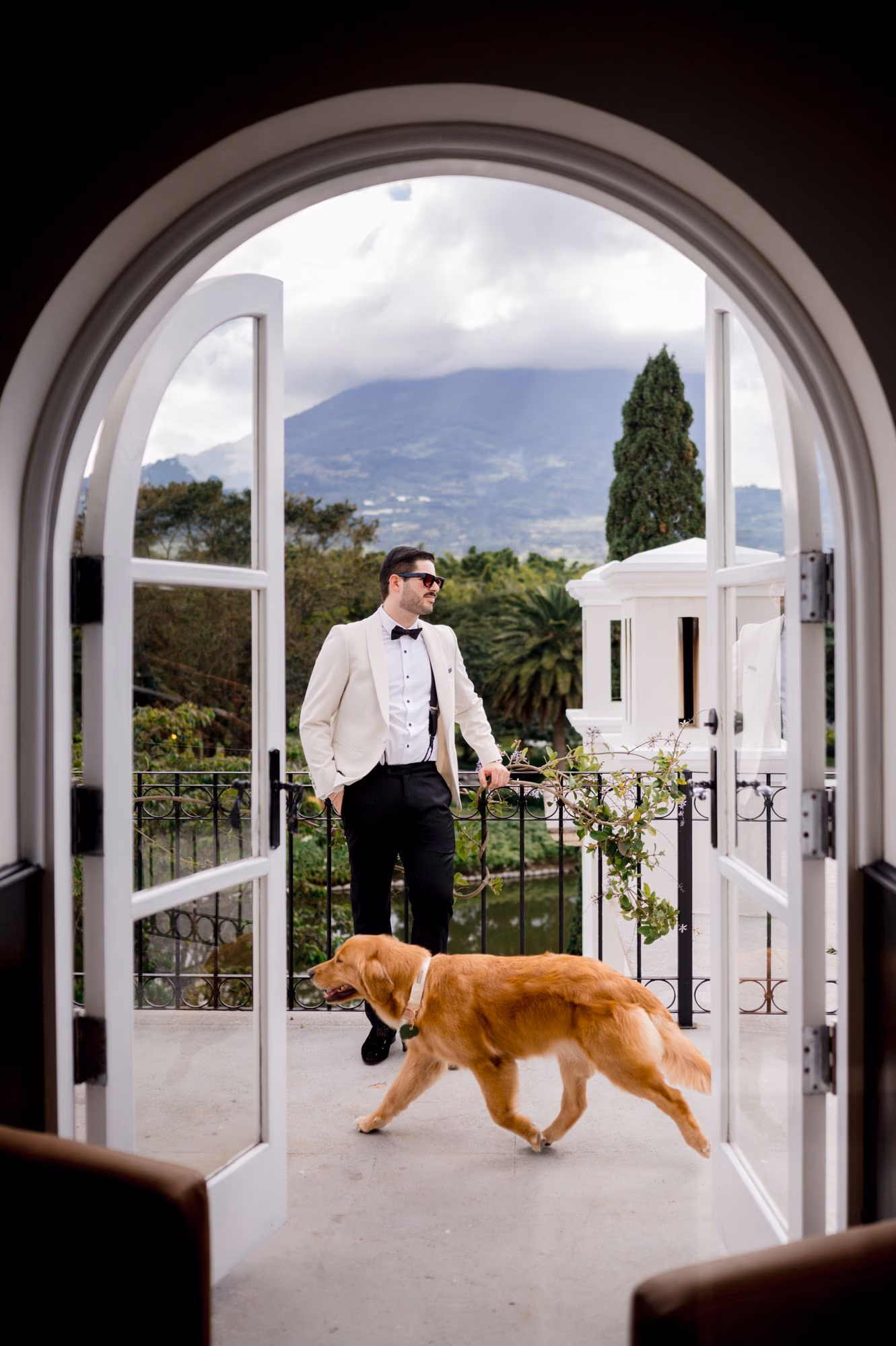 Sergio waits on a terrace framed by arched doors as Nala the golden retriever trots through — La Antigua Guatemala wedding morning.