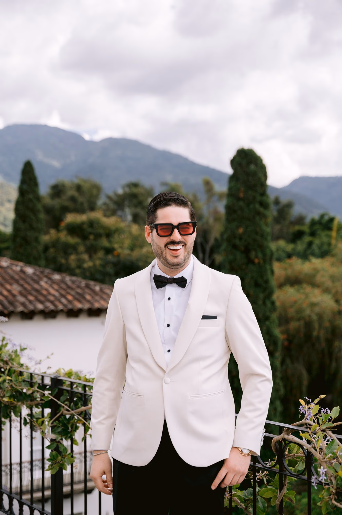 Groom laughs in a white tuxedo and red-tinted sunglasses with mountains behind him at Villa Bokeh.