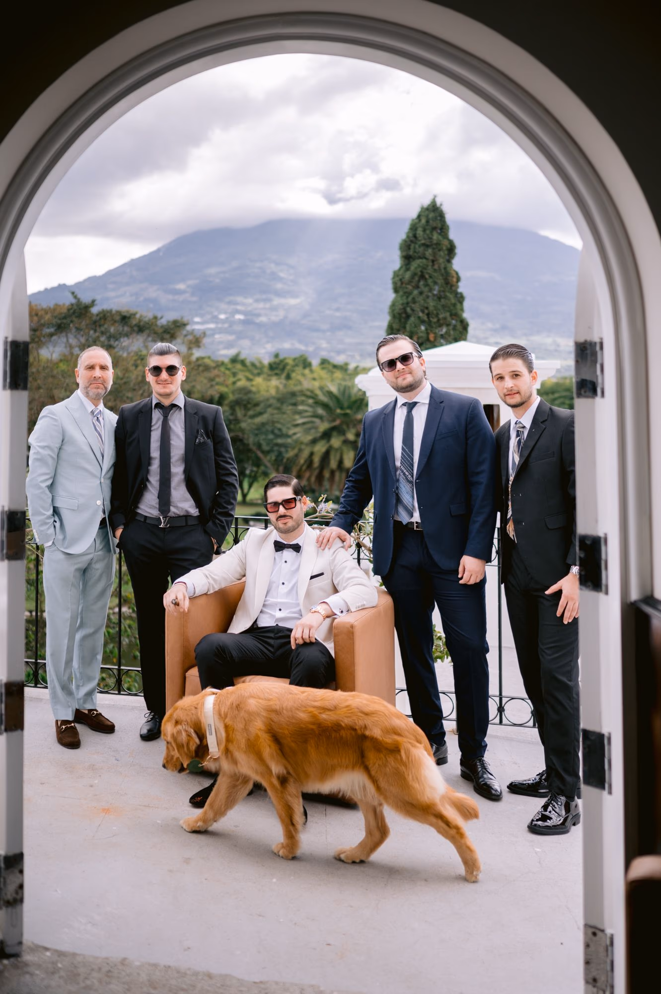Groomsmen surround Sergio seated in a leather armchair while Nala the golden retriever walks past — destination wedding in Antigua Guatemala.