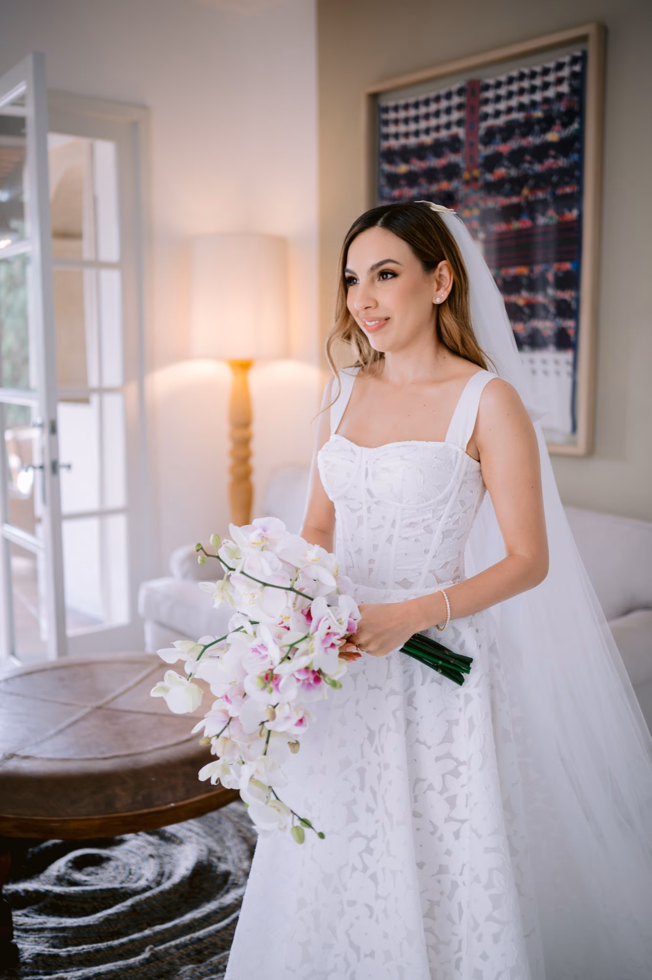 Caro stands by a lamplit window in her lace gown holding a cascading orchid bouquet — bridal portrait at Villa Bokeh.