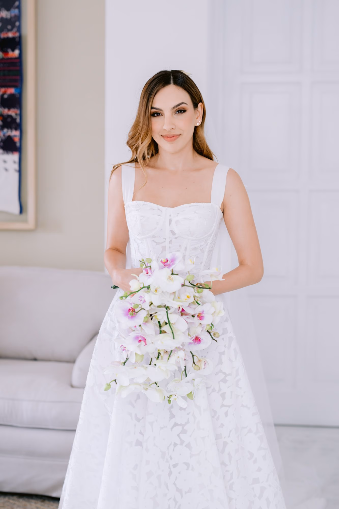 Caro smiles softly holding a cascading orchid bouquet in a bright getting-ready suite — intimate bridal portrait.