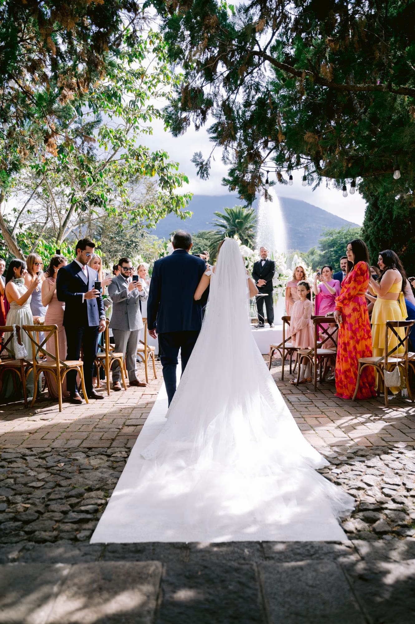 Bride walks down the cobblestone aisle with her father, cathedral veil trailing behind toward the fountain altar.