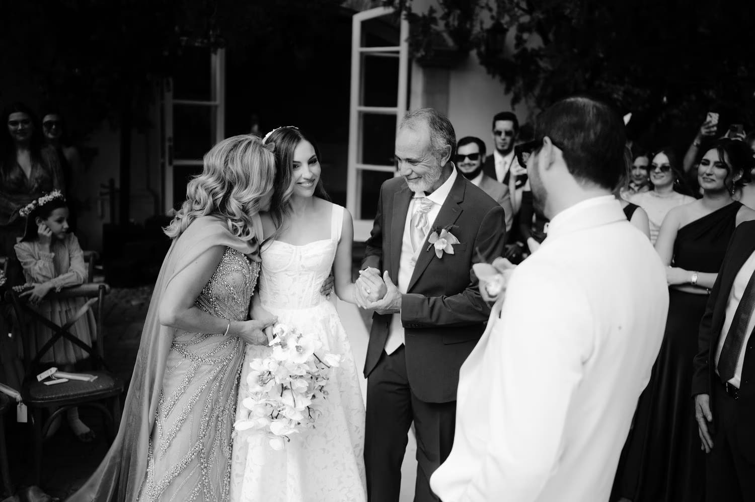 Bride embraces her mother at the head of the aisle while her father waits beside her — emotional ceremony moment in black and white.