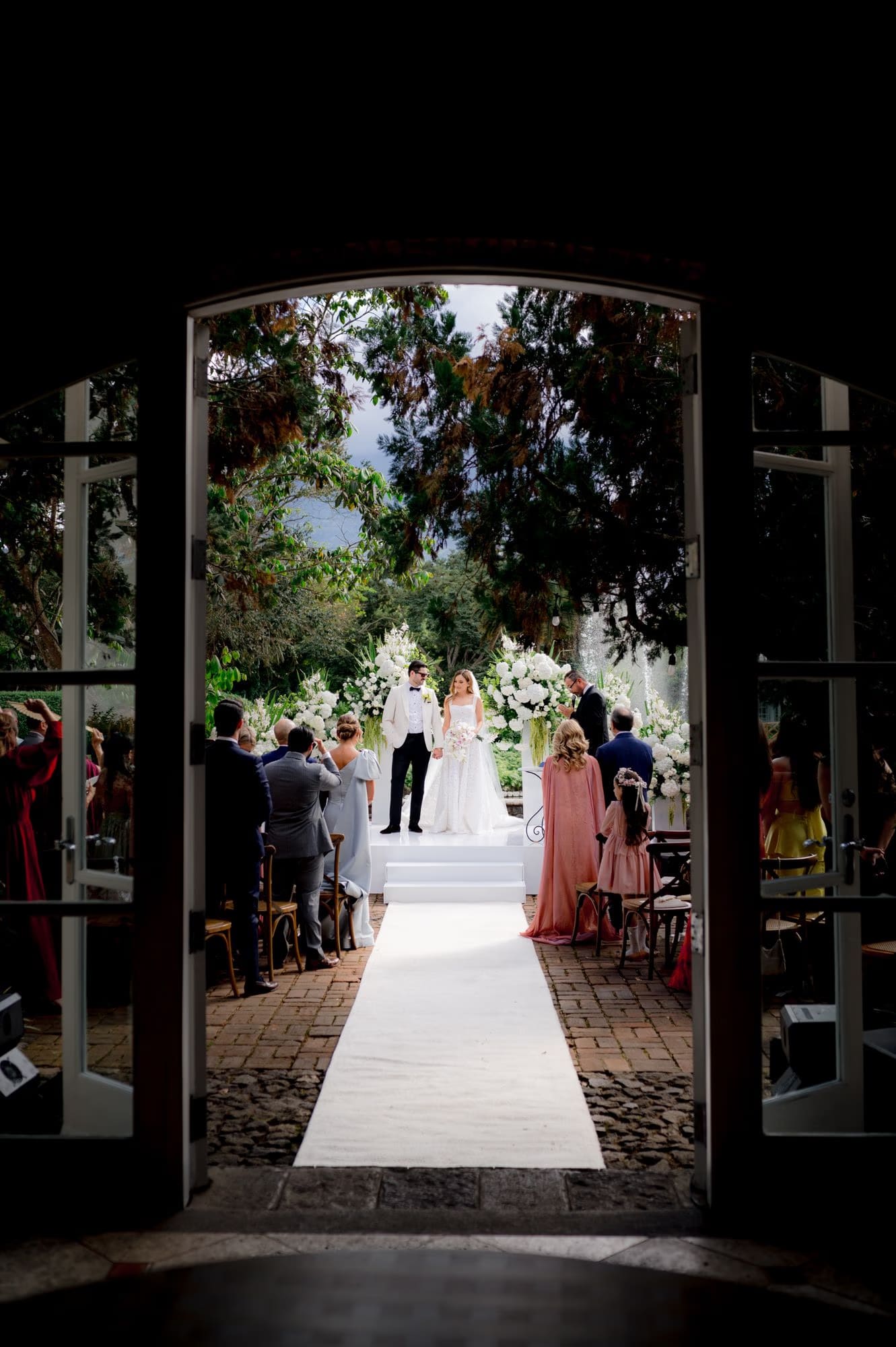 Ceremony framed through a dark archway, aisle leading to bride and groom in the sunlit garden beyond.