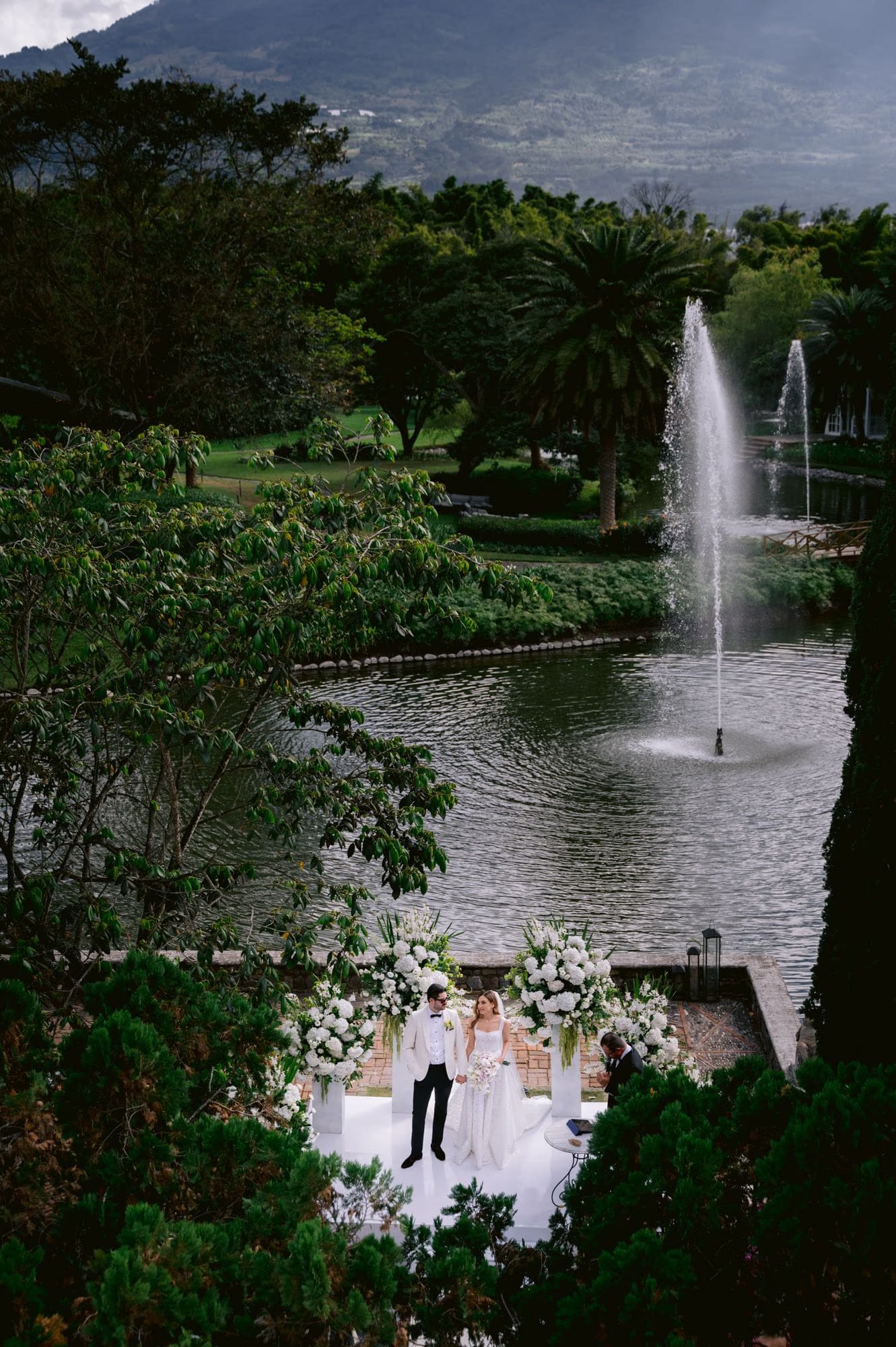 Aerial of the couple at their lagoon altar with the towering fountain and volcano beyond at Villa Bokeh.