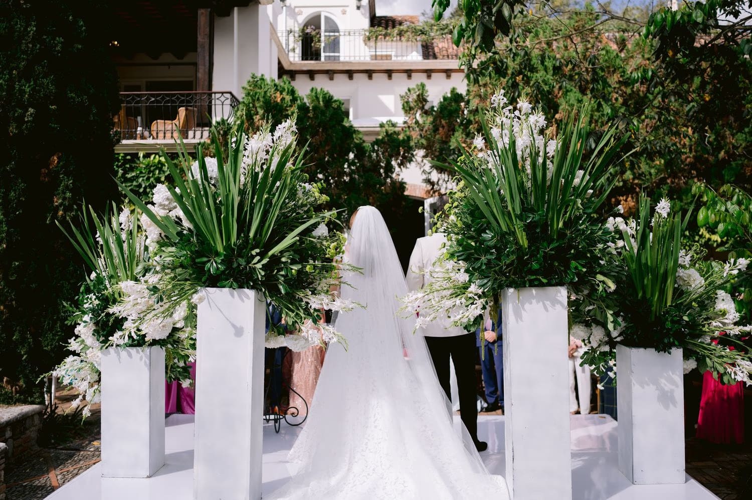 Bride walks between tall white floral pedestals toward the colonial facade at Villa Bokeh.
