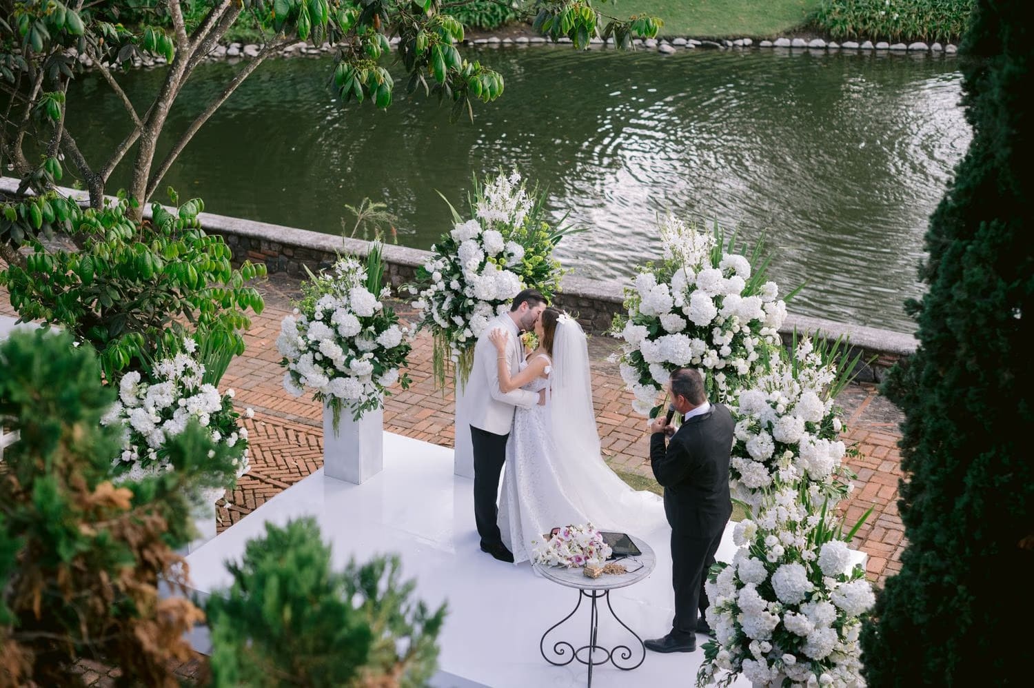 Aerial first kiss between white floral pillars by the lagoon — wedding ceremony at Villa Bokeh.