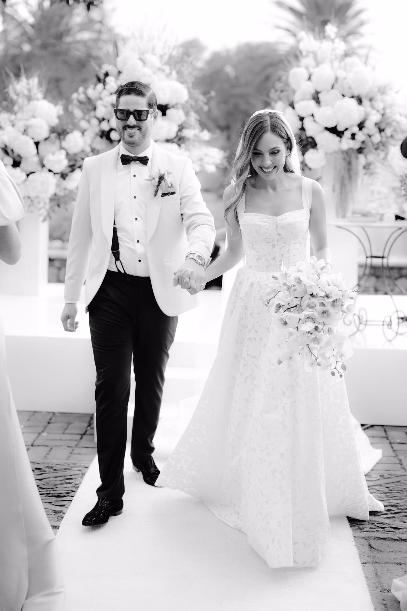 Newlyweds walk back down the aisle holding hands, laughter on their faces, in black and white.