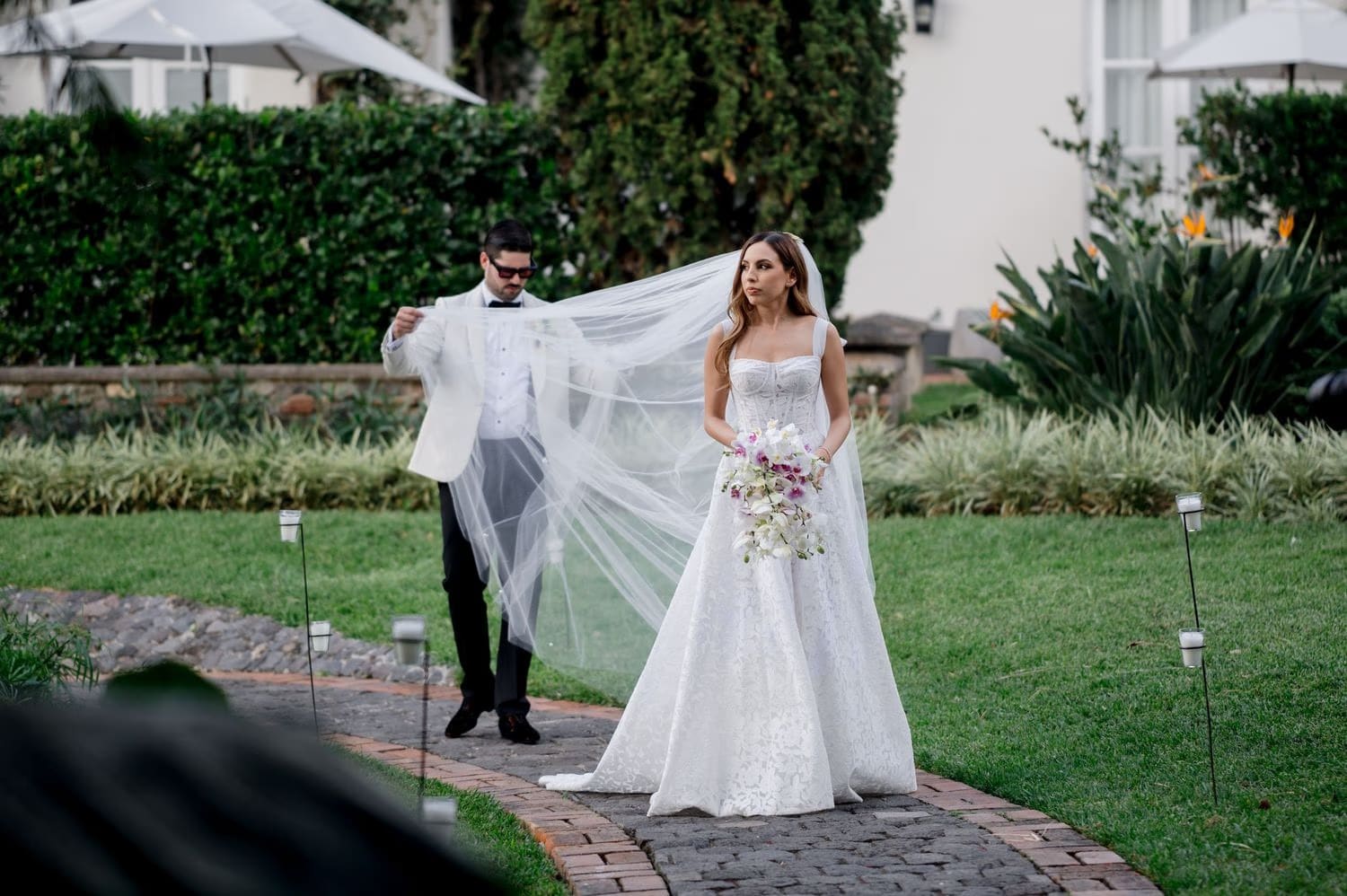 Groom adjusts Caro's veil on a garden path while she holds her orchid bouquet — Villa Bokeh portraits.