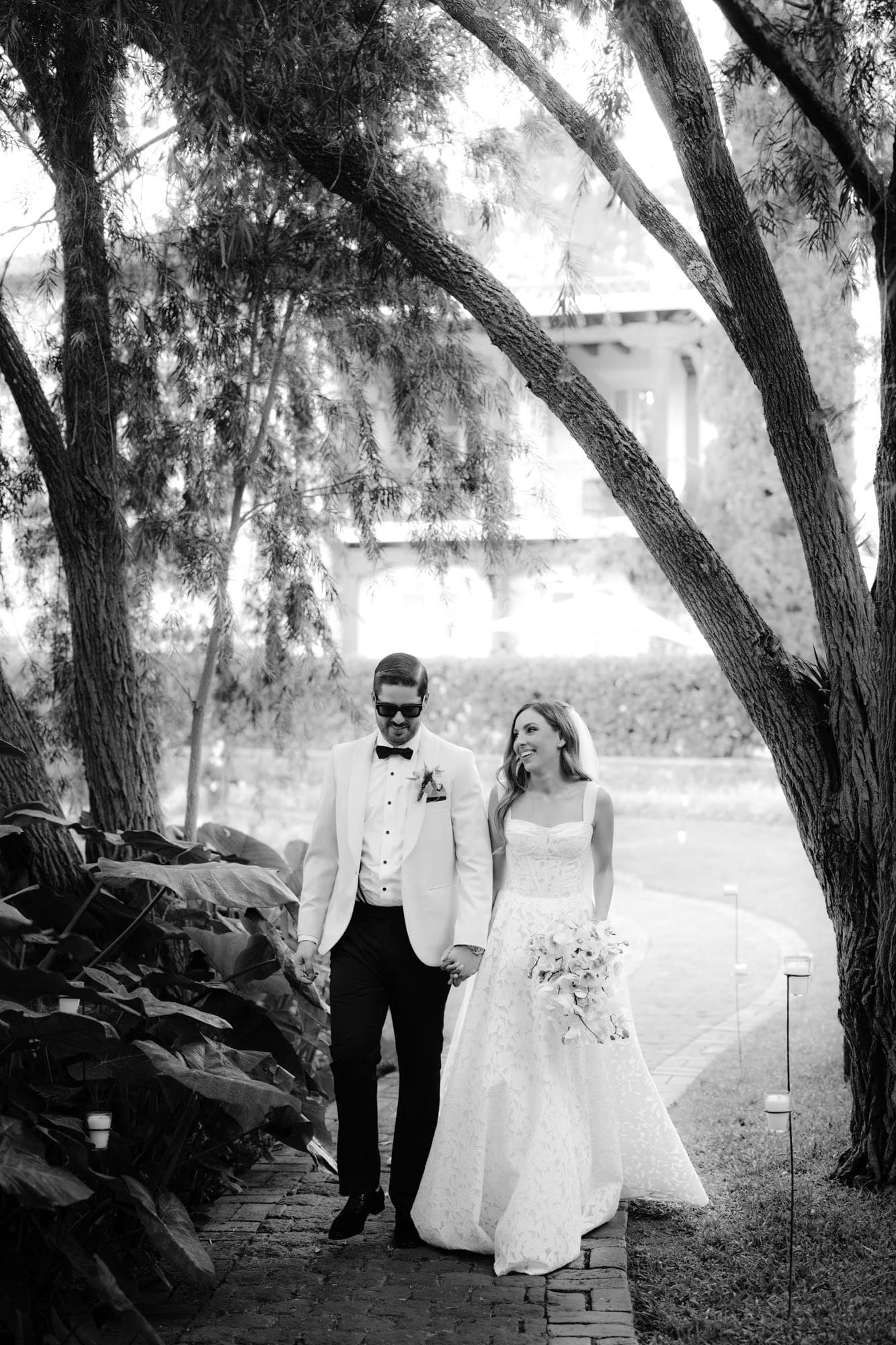 Bride and groom walk under arching trees in black and white — editorial portrait in Antigua.