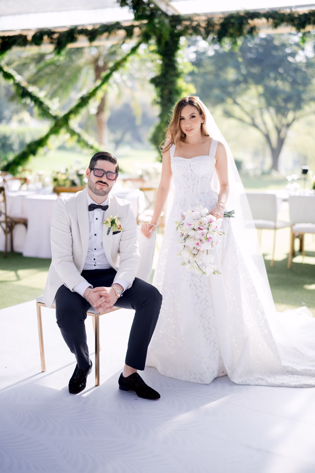 Sergio seated beside Caro standing under the glass tent reception canopy — Villa Bokeh portrait.