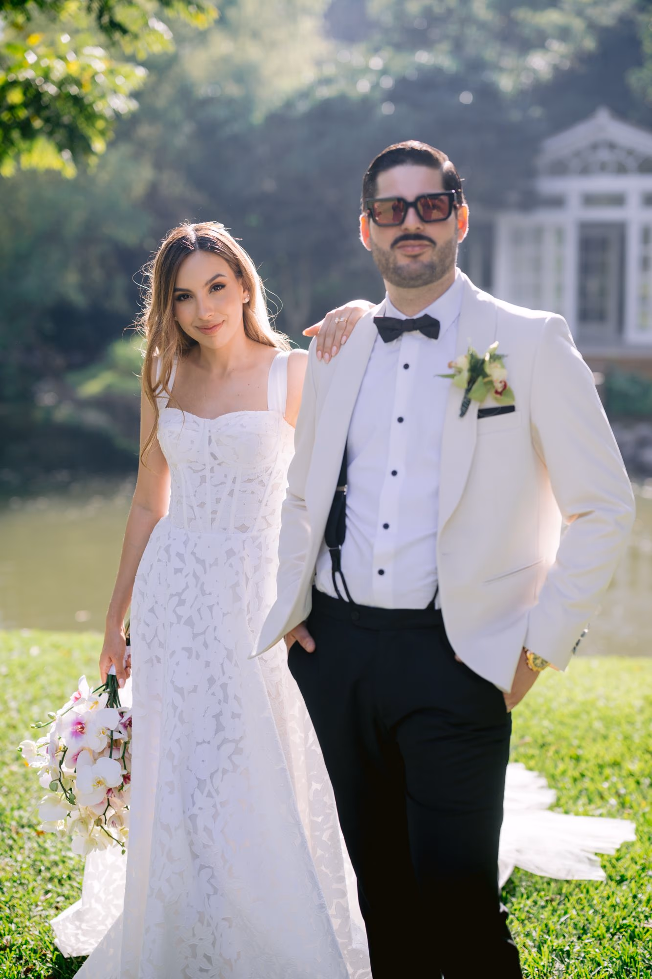 Bride and groom share a quiet smile beside the lagoon at Villa Bokeh, garden house behind them.