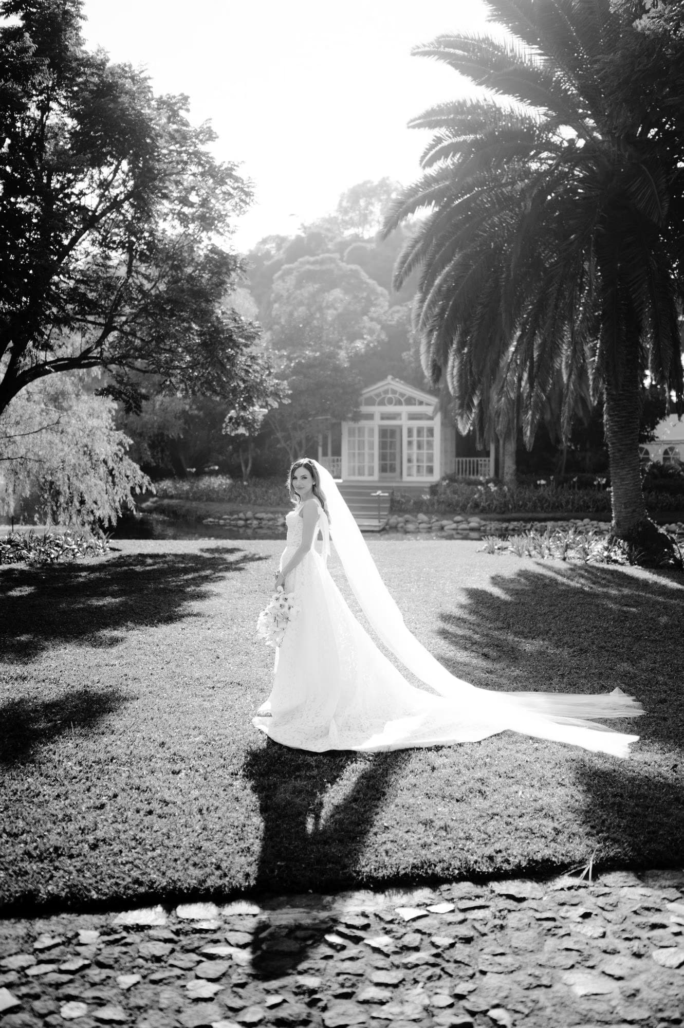 Black-and-white bridal portrait, veil and dress trail spreading across the lawn.