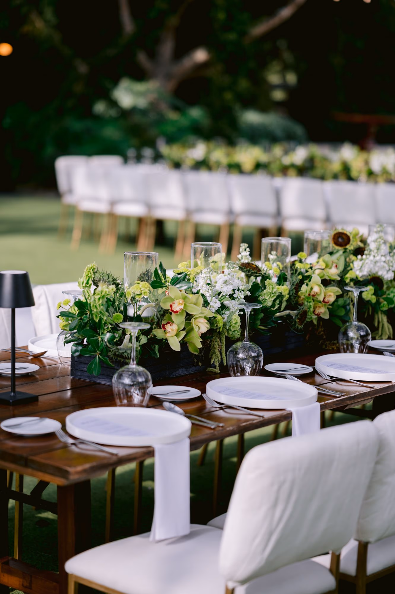 Long farm table set with green floral runner and white chairs at Villa Bokeh reception.