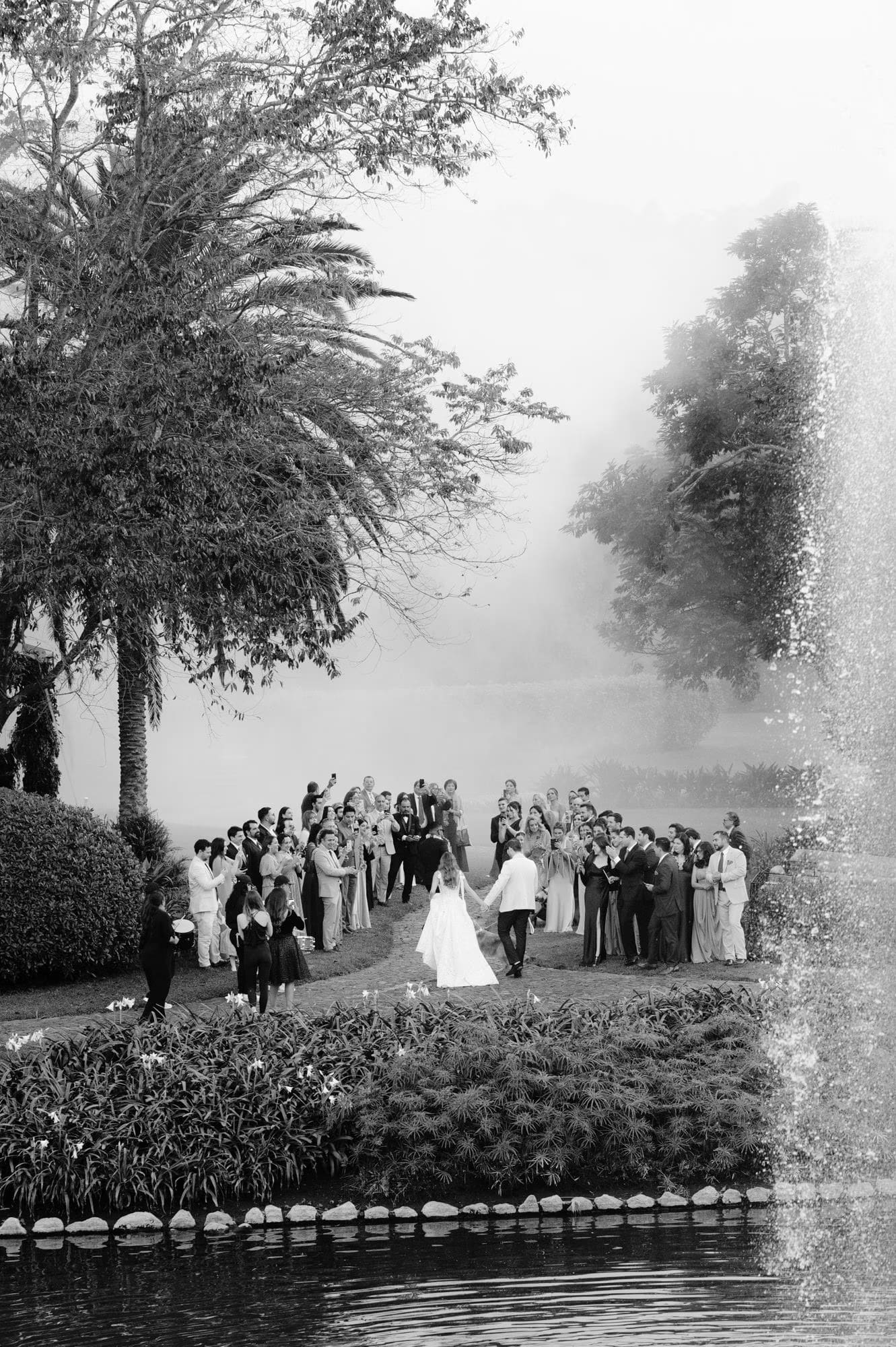 Black-and-white view of guests cheering as the newlyweds walk past the fountain at Villa Bokeh.