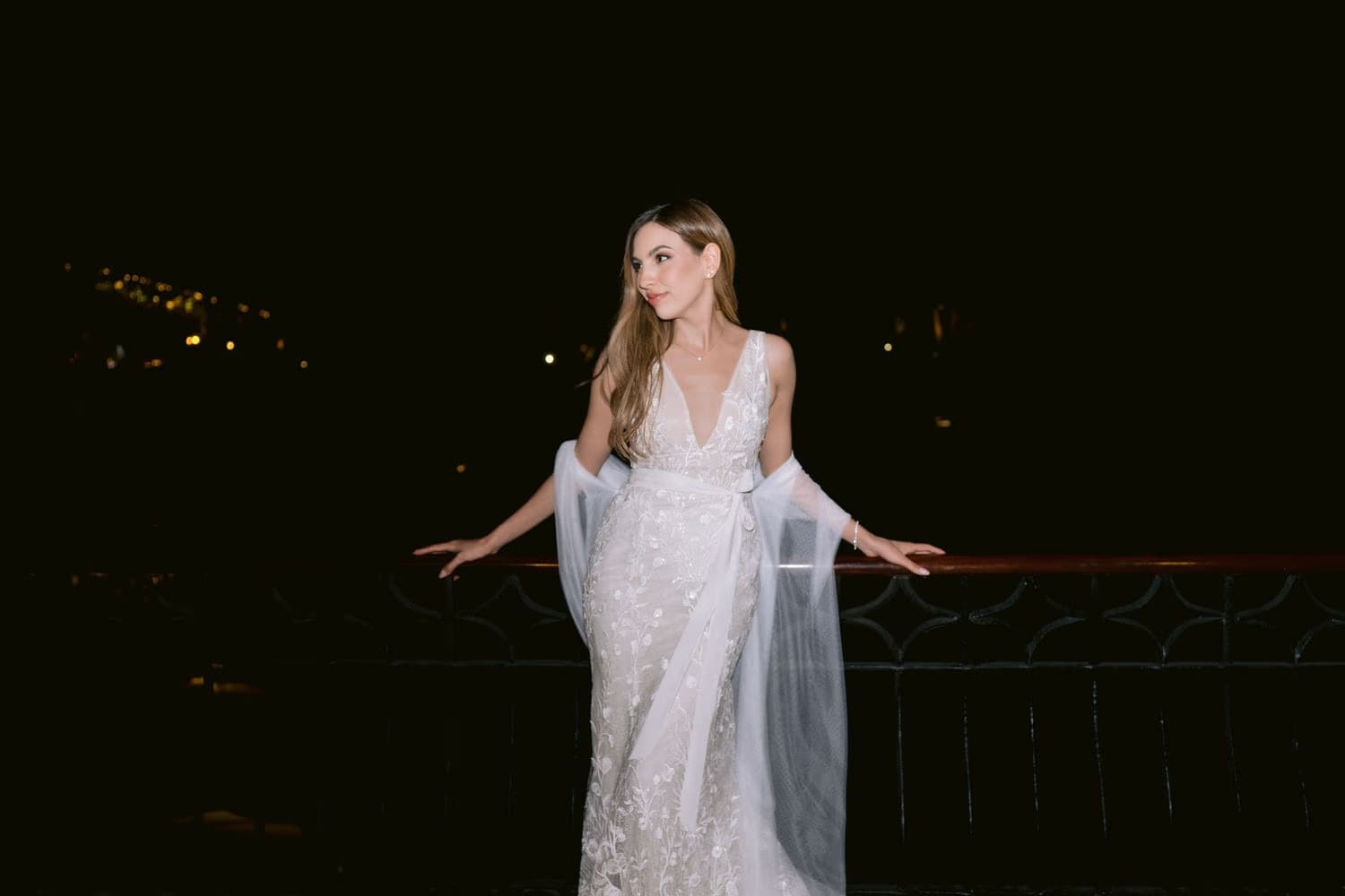 Bride leans on a wrought-iron rail at night, embroidered second-look dress catching the light.