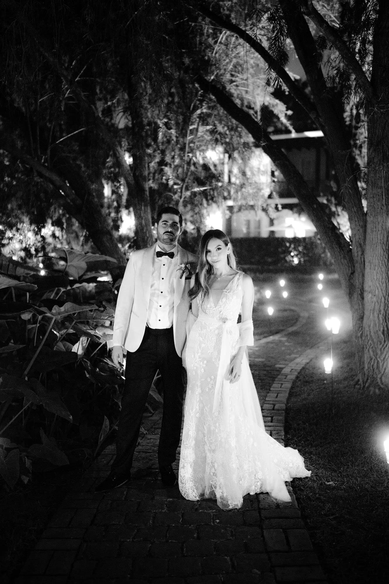 Newlyweds stand on a candlelit garden path at night in black and white — Villa Bokeh reception.