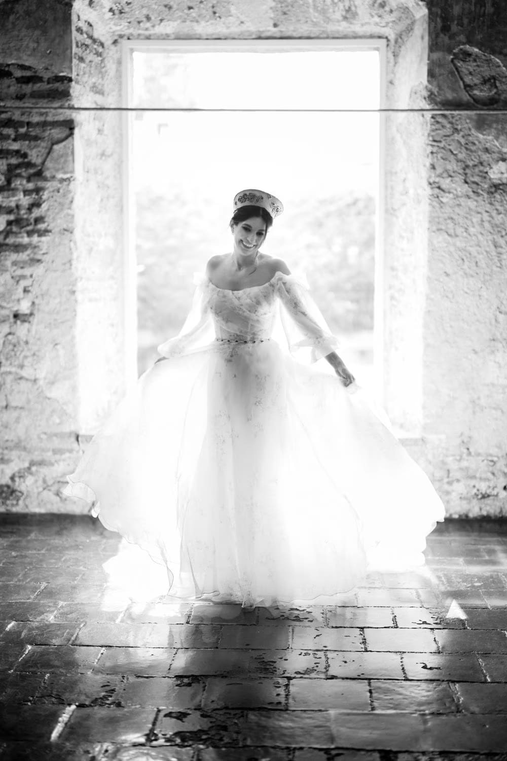 Bride twirls in floral gown inside a stone-walled archway with backlit window — black-and-white bridal portrait in Antigua Guatemala.