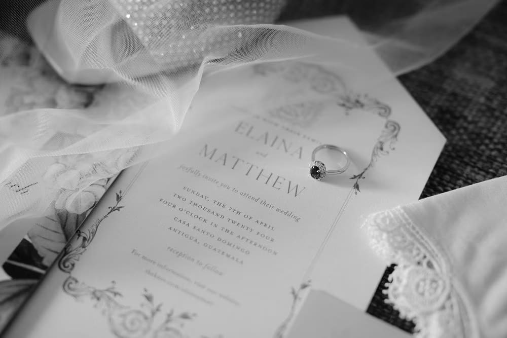 Black-and-white close-up of engagement ring on engraved wedding invitation framed by tulle veil — bridal preparation detail.