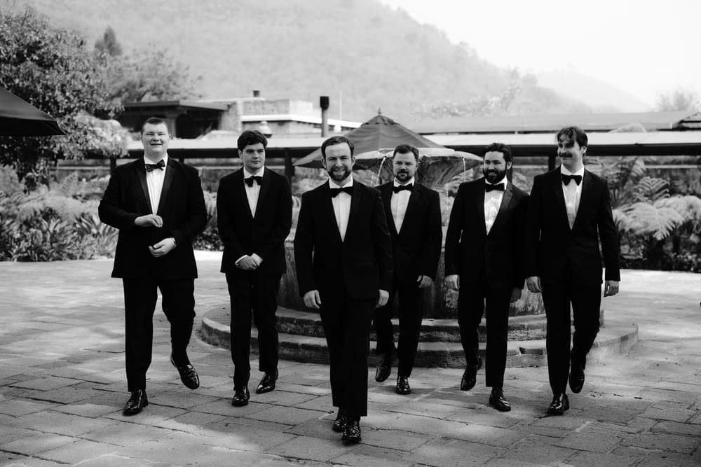 Groom Matt walks with his groomsmen in matching tuxedos past a volcano-view fountain — black-and-white destination wedding portrait in Antigua.