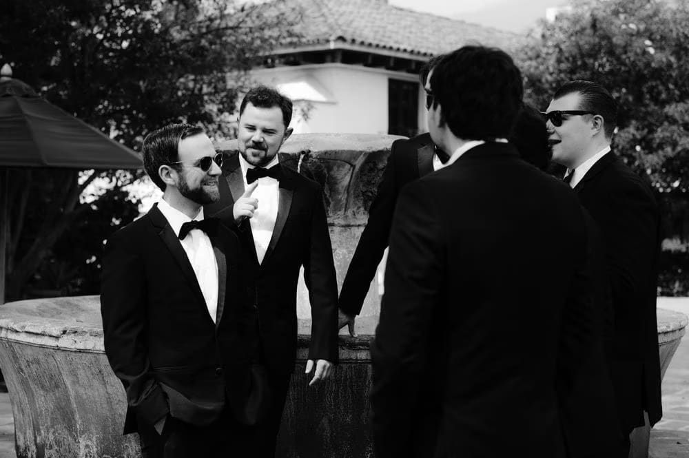 Groomsmen share a laugh around a stone fountain before the ceremony in colonial Antigua Guatemala — candid black-and-white moment.