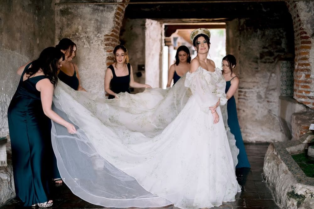 Bridesmaids in navy gowns adjust the bride's flowing train inside a stone corridor — getting-ready moment at the restored monastery.