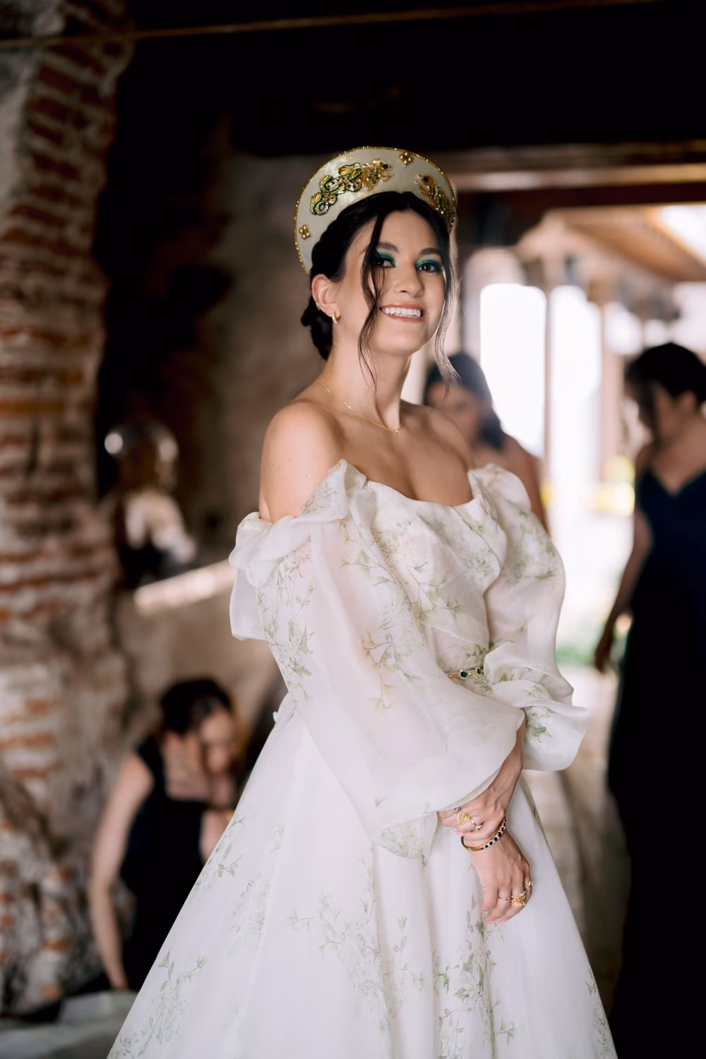Bride smiles in beaded crown and floral gown under a brick archway during preparation — destination bridal portrait.