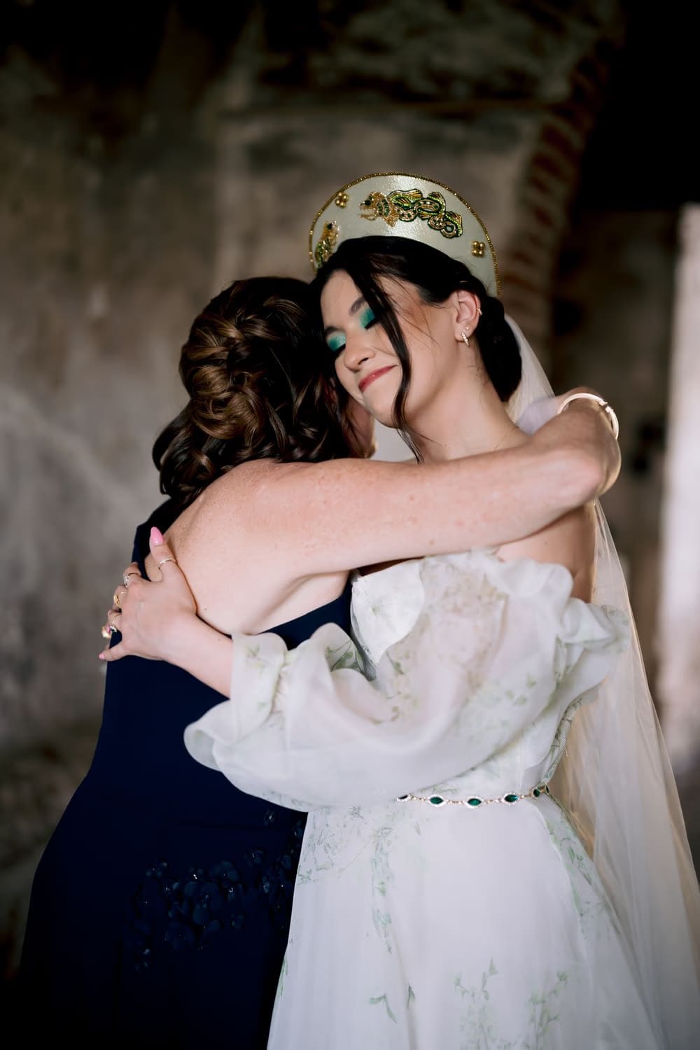 Bride hugs her mother with eyes closed, beaded headpiece in focus — emotional bridal preparation moment.