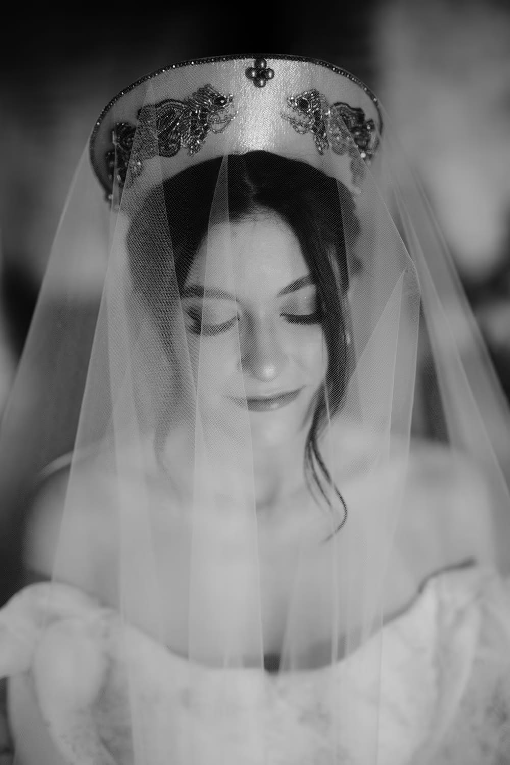Black-and-white bridal portrait under a beaded headpiece veil with eyes lowered — editorial getting-ready moment.