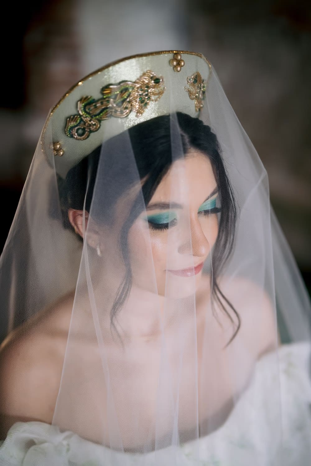 Bride glances down beneath embroidered crown and sheer veil with emerald-green eyeshadow — close-up bridal portrait.