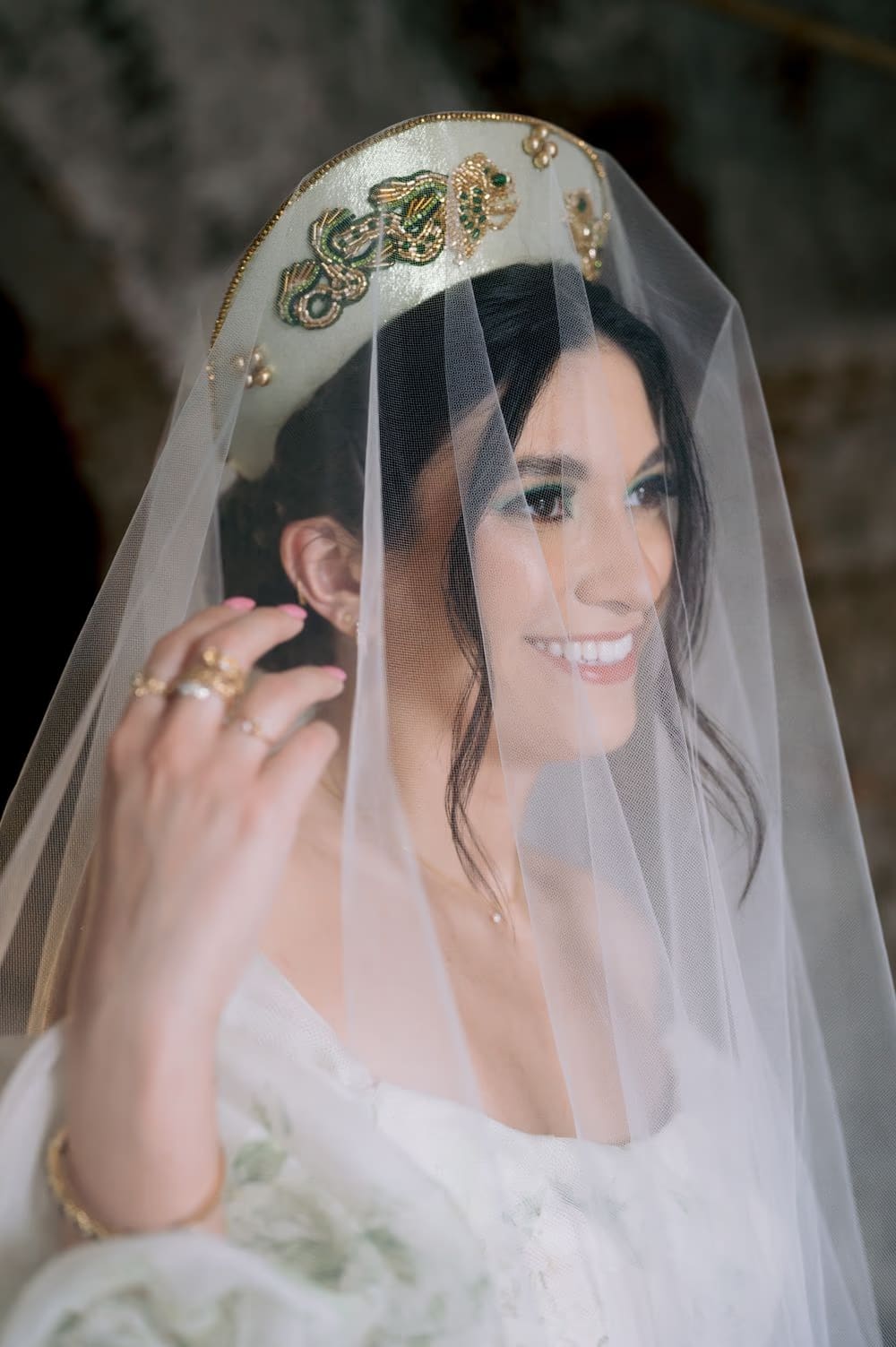 Bride lifts her veil with a wide smile, showing stacked gold rings and beaded crown — radiant getting-ready portrait.