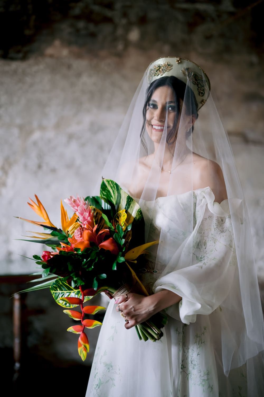 Bride holds vibrant tropical bouquet of heliconia and ginger under stone arches — destination bridal portrait.