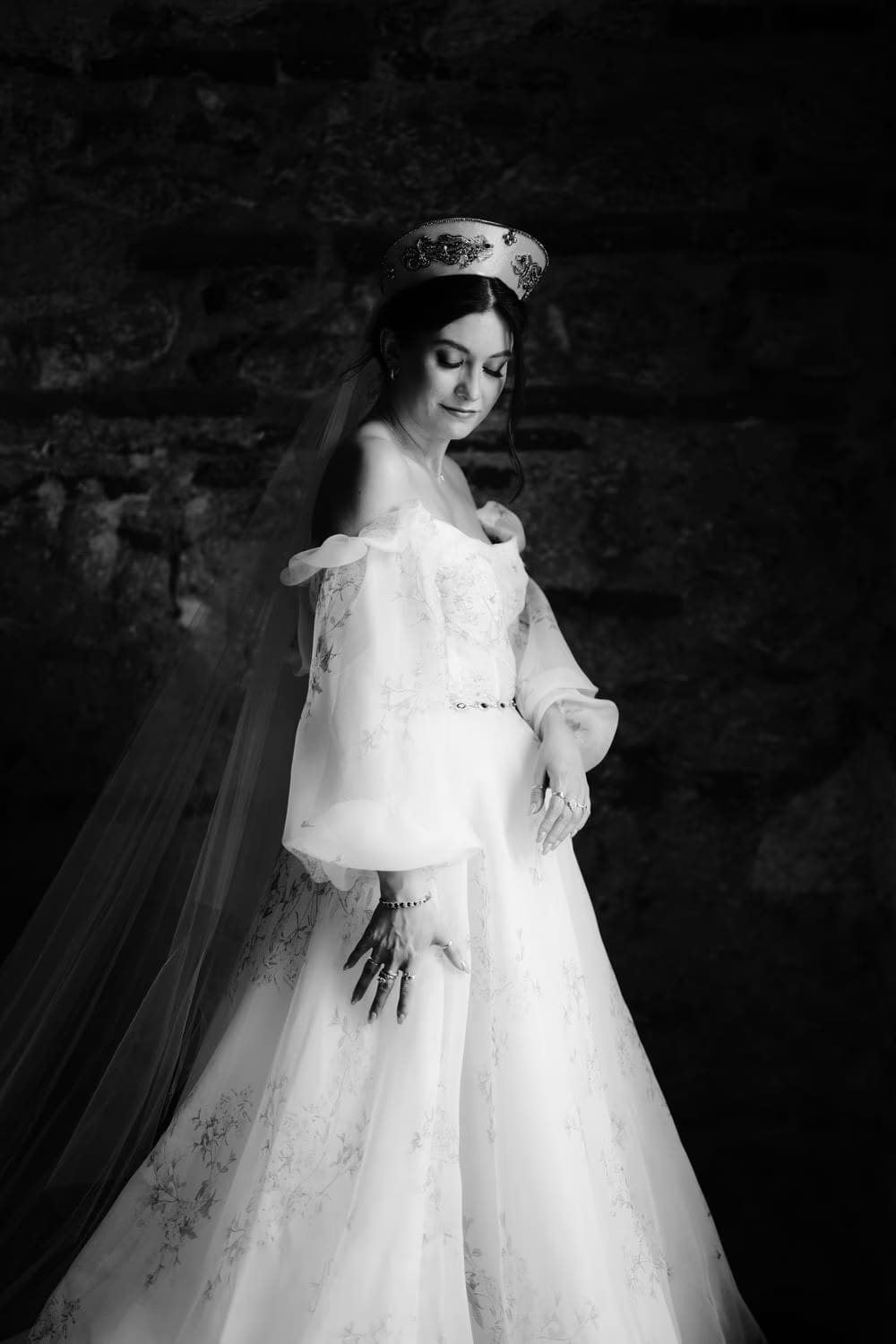 Black-and-white full-length bridal portrait against a dark stone wall, showing the cathedral train and beaded crown.