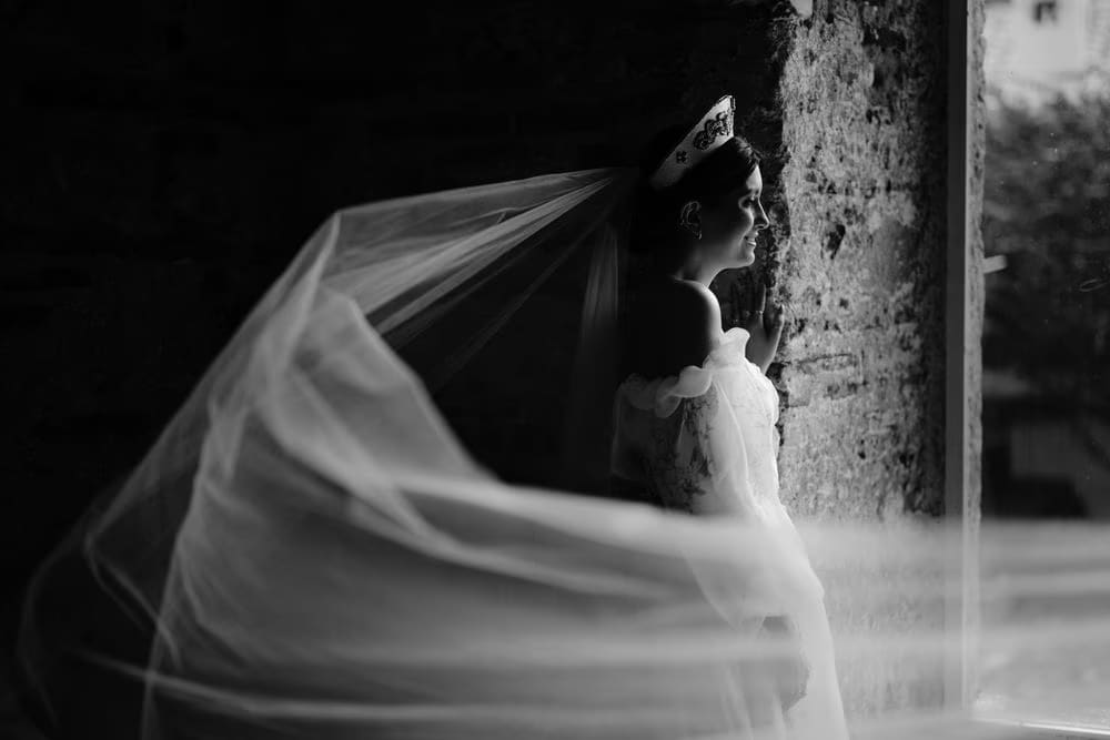Bride's veil drifts in the breeze against a weathered stone wall — cinematic black-and-white profile.