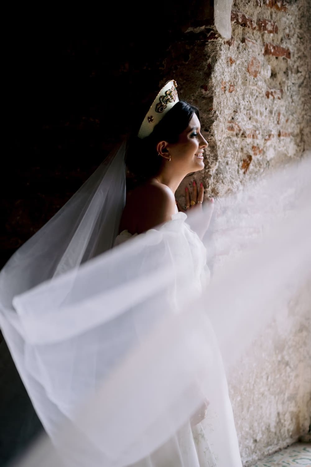 Bride leans against a brick wall as her veil catches sidelight — editorial profile portrait.