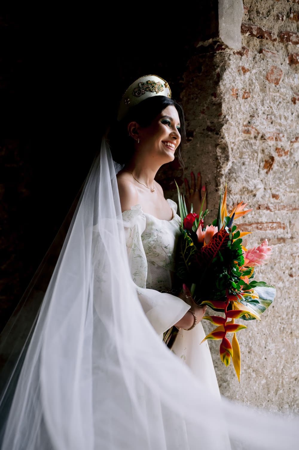 Bride laughs holding her tropical bouquet beside a sunlit brick wall — radiant bridal portrait.