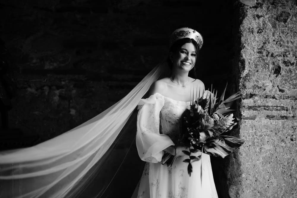 Black-and-white bridal portrait with cascading veil and tropical bouquet beside textured stone wall.