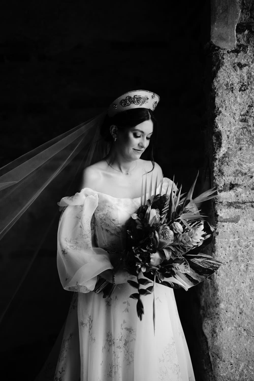 Bride looks downward at her bouquet of proteas and palms in black-and-white — moody editorial portrait.