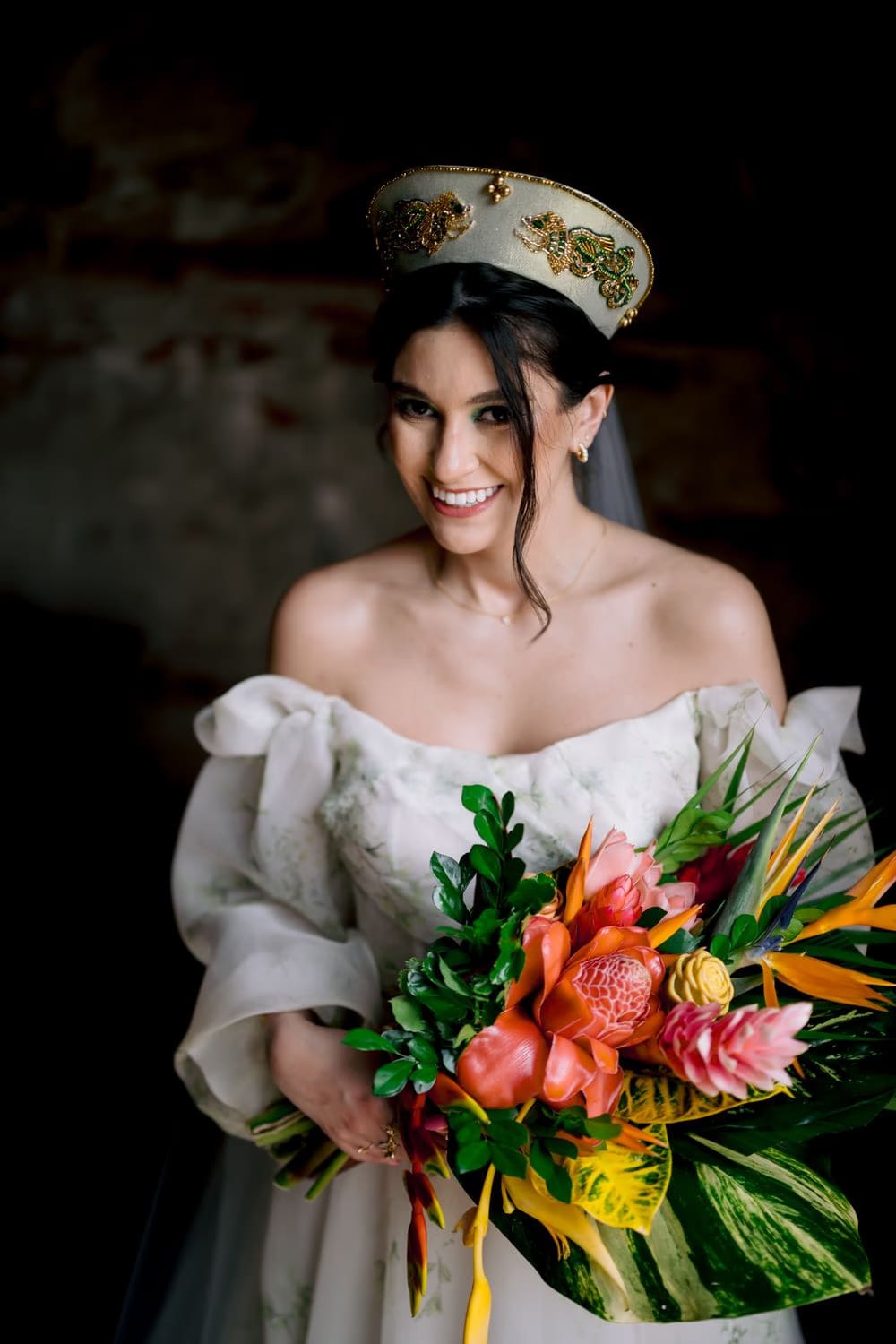 Smiling bride holds a wide tropical bouquet of ginger, croton and birds of paradise against a dark stone wall.