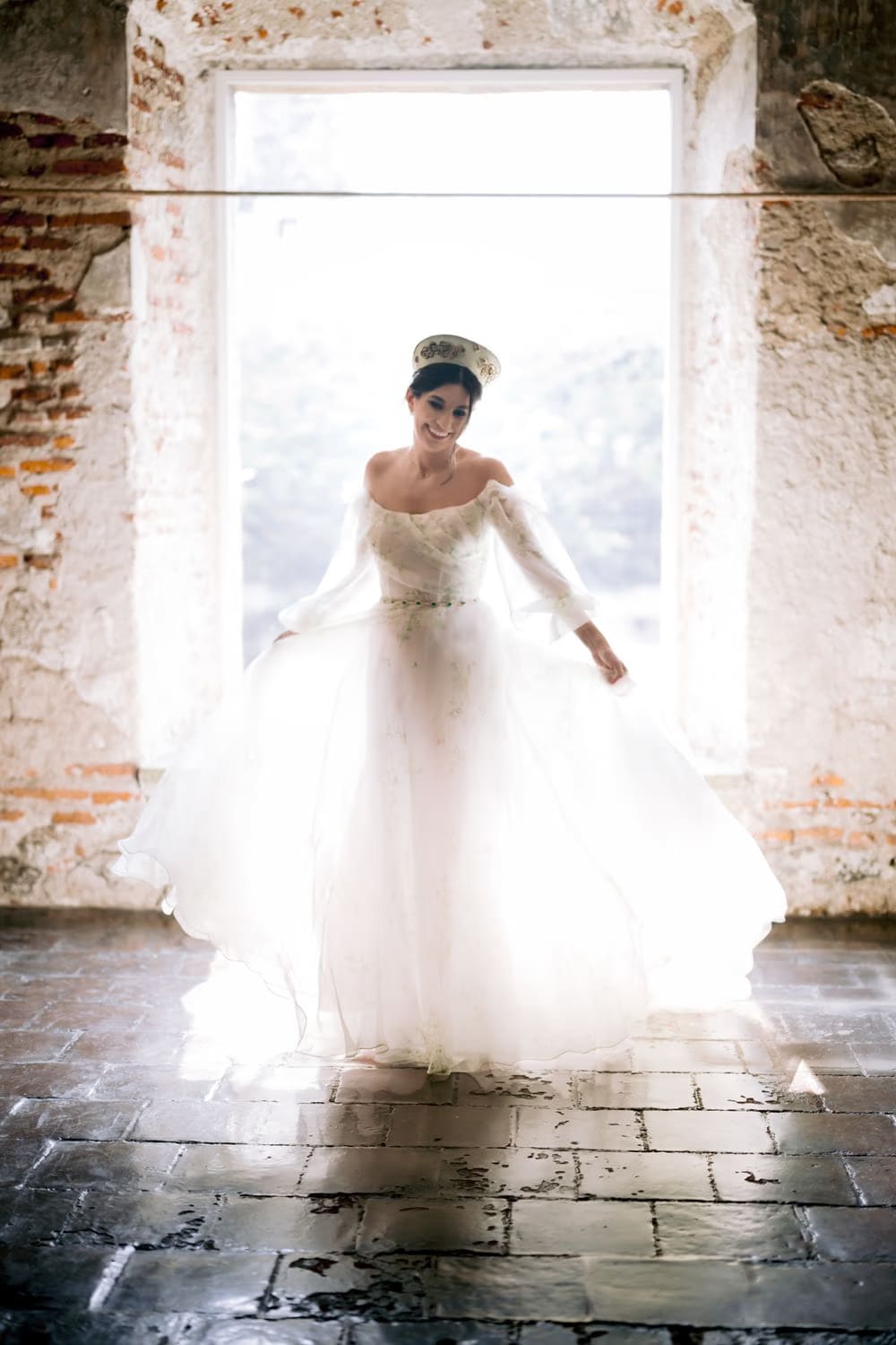 Bride twirls her gown inside a brick-framed window, light flooding the stone floor — destination bridal portrait.