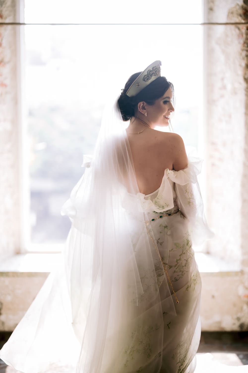 Back view of the bride at a tall window, veil flowing and floral gown catching morning light.
