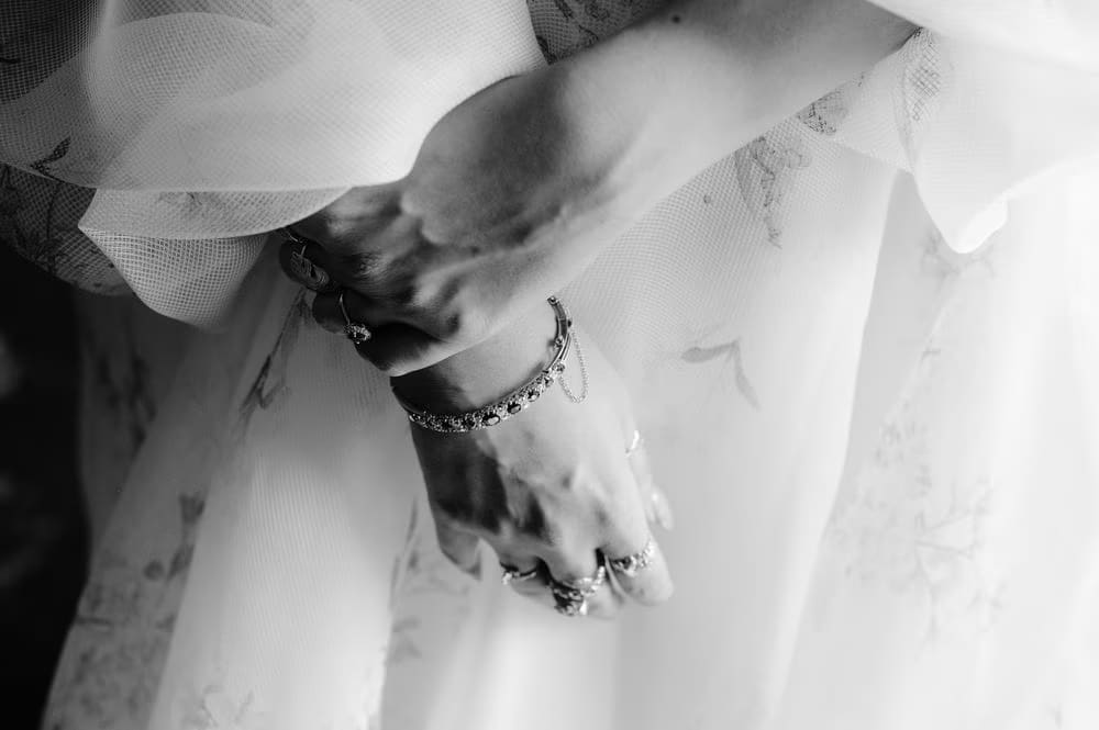 Black-and-white close-up of the bride's hands with diamond bracelet and stacked rings over tulle.