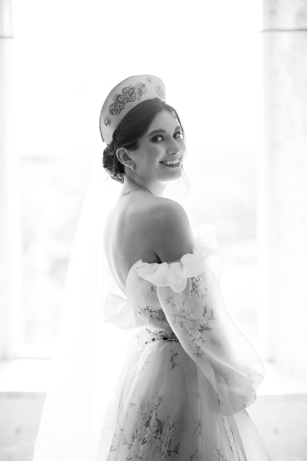 Black-and-white bridal portrait glancing back over a sheer floral sleeve against bright window light.