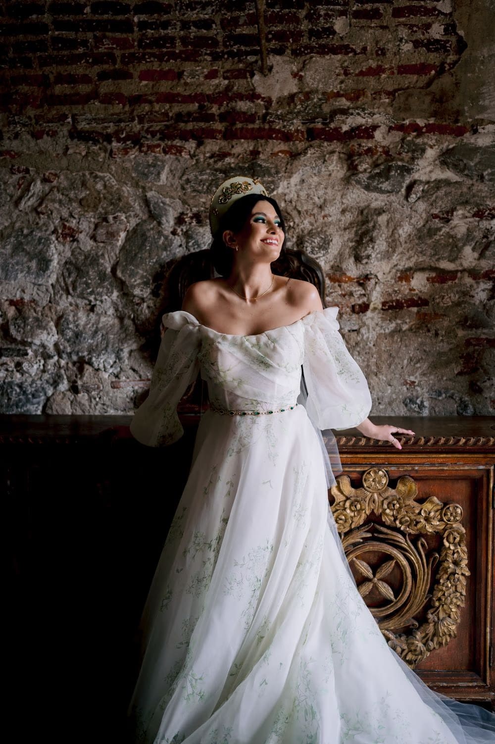 Bride leans on a carved wooden bench against rough stone wall — editorial portrait beside antique church furniture.