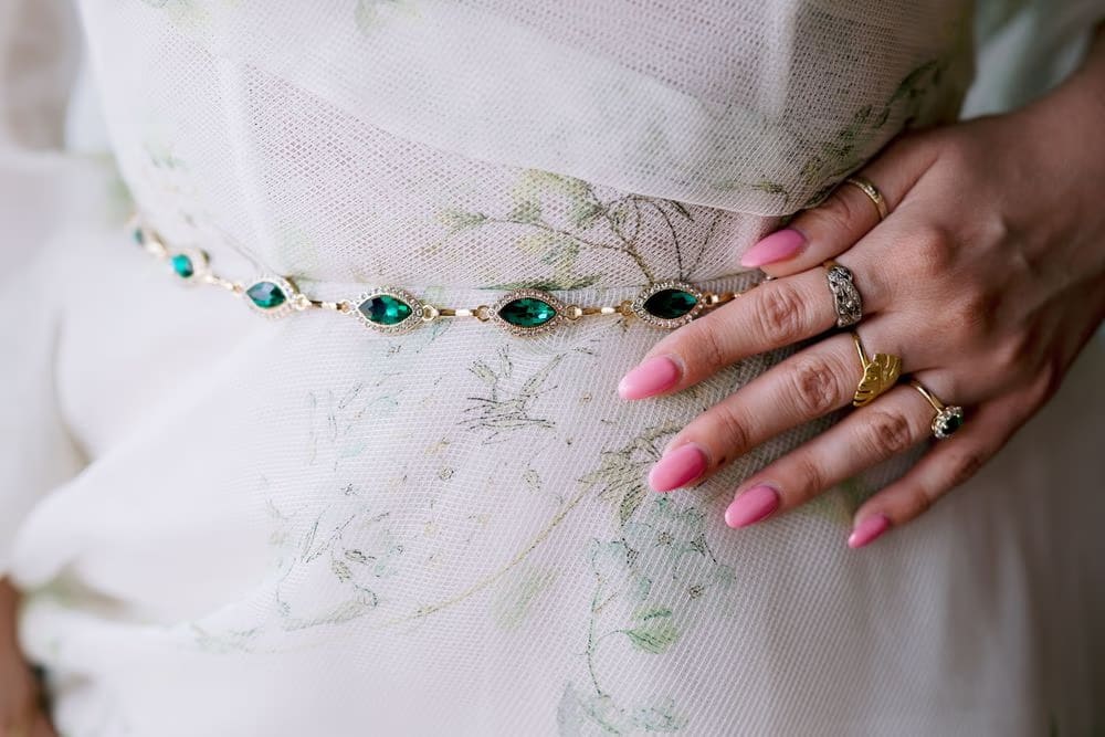 Close-up of emerald belt and stacked sapphire and gold rings against floral organza gown — bridal jewelry detail.